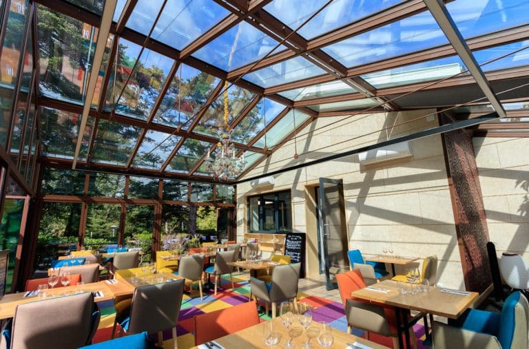 Une pergola en aluminium, peinte en marron, s'adosse à une maison. Le paysage de forêt autour, créé un cadre verdoyant. Une table et des chaises disposées sous la pergola invitent à la détente dans un environnement paisible.