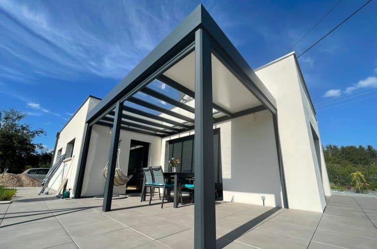 Une pergola en aluminium, s'adosse à une maison. Une table et des chaises disposées sous la pergola invitent à la détente. Le soleil brille et le beau temps incite à profiter du calme de ce petit coin de paradis.